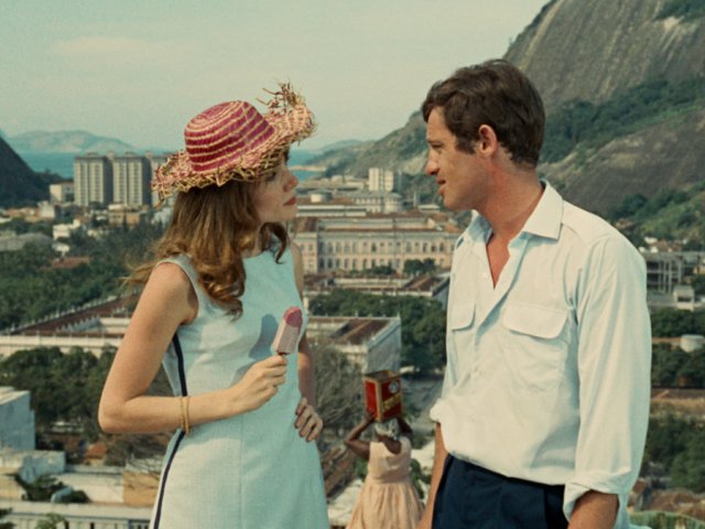 photogramme du film L'Homme de Rio montrant un personnage féminin à gauche, joué par Françoise Dorléac, en robe bleue avec un chapeau de paille, tenant une glace à la main et regardant un personnage masculin à droite, joué par Jean-Paul Belmondo, habillé d'une chemise blanche. Il s'agit d'un plan rapproché taille avec la ville de Rio en arrière-plan.